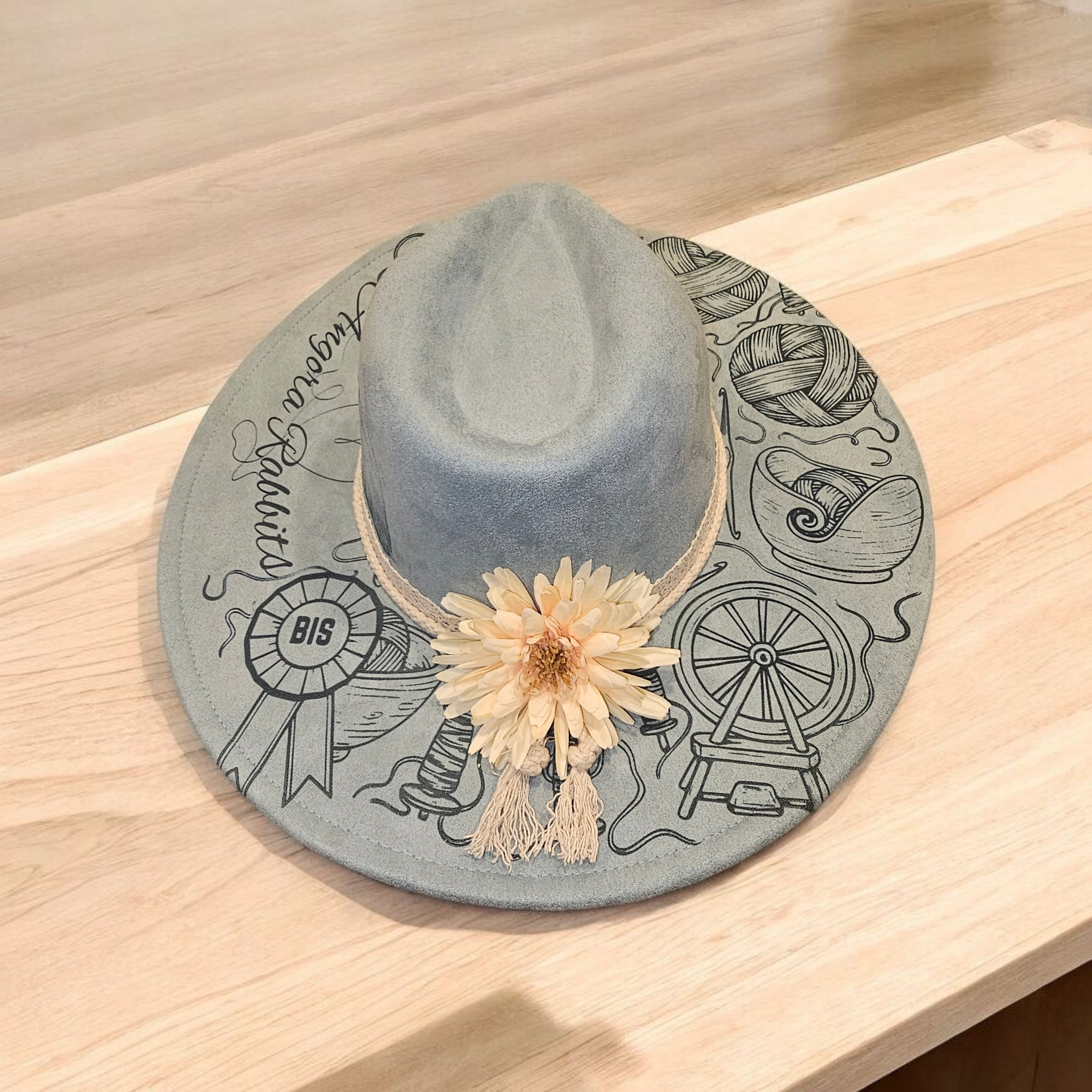 Gray hat with Angora Specialty wording, BIS rosette, yarn and spinning-wheel artwork, and a pale flower